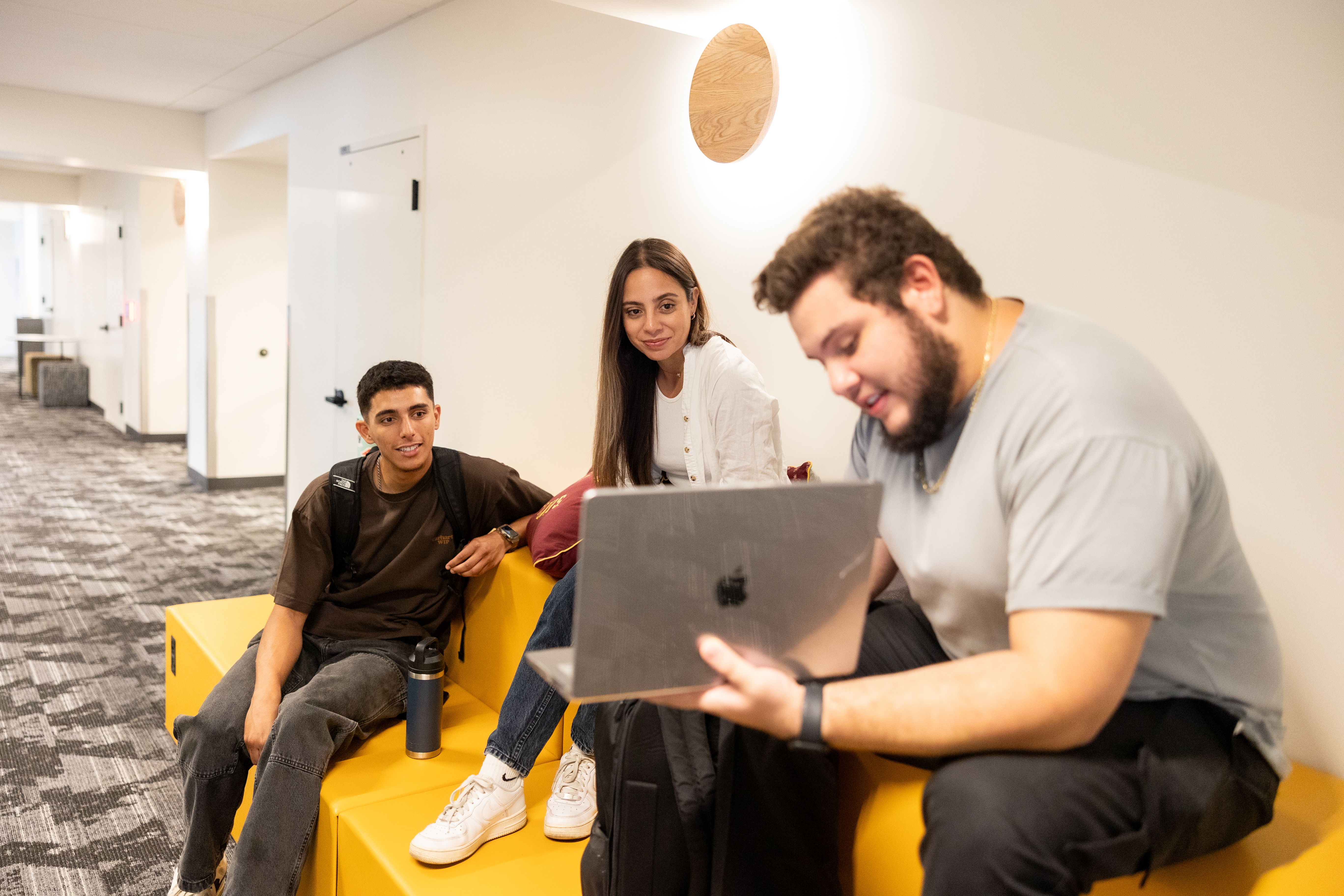 Three young adults seated on yellow modular seating in a corridor, looking at a laptop together.