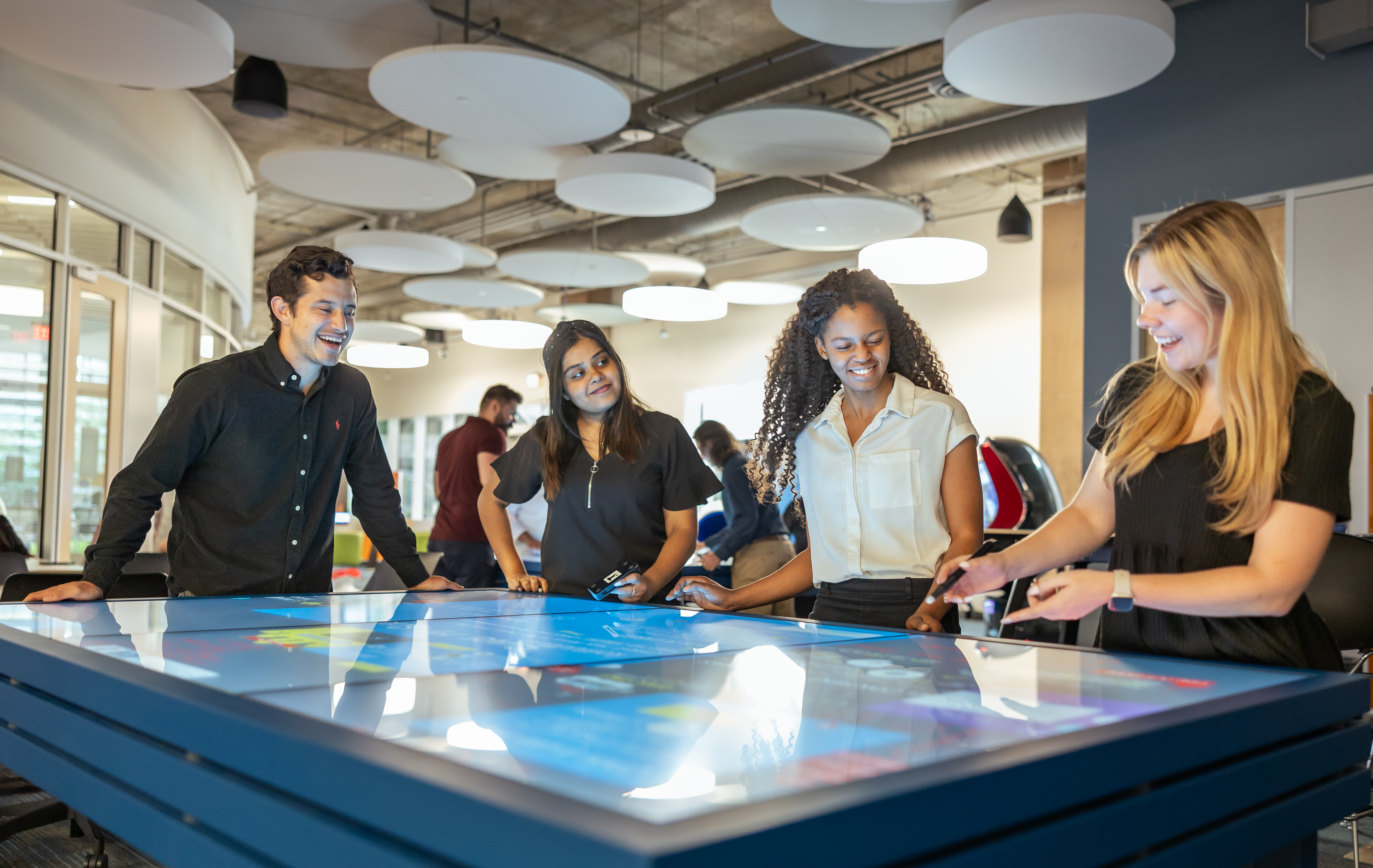 Four people interacting with a large touchscreen table in a modern, brightly lit room.