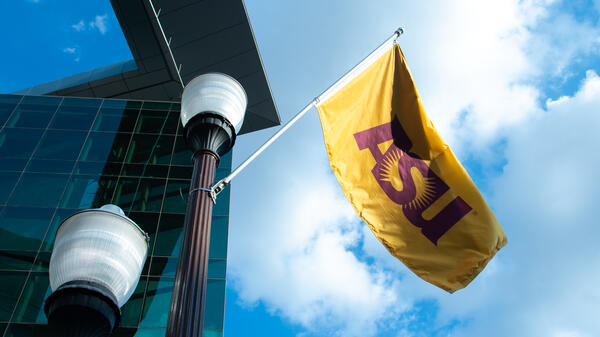 A gold flag flying near streetlights and a modern building with blue-tinted windows under a partly cloudy sky.